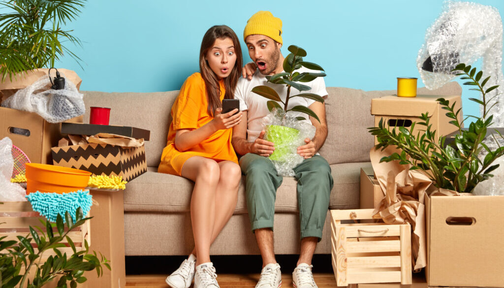 Emotive young woman and man stare at smartphone device, search new design for living room, relocate in new abode, hold potted plant, real mess with carton boxes around. Young homeowners on sofa