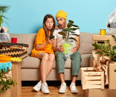 Emotive young woman and man stare at smartphone device, search new design for living room, relocate in new abode, hold potted plant, real mess with carton boxes around. Young homeowners on sofa